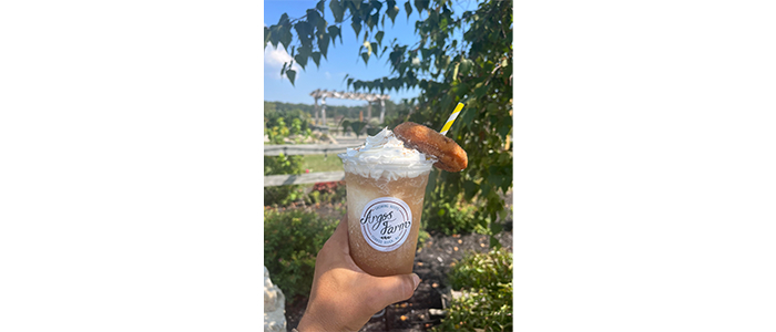 person holding a cold drink with whipped cream, straw and a donut overlooking a field