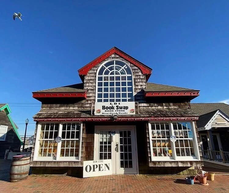 Exterior photo of LBI Book Swap with a blue sky, a seagull flying. The exterior is wood with white windows and red trim around the roof with a sign that says book store open.