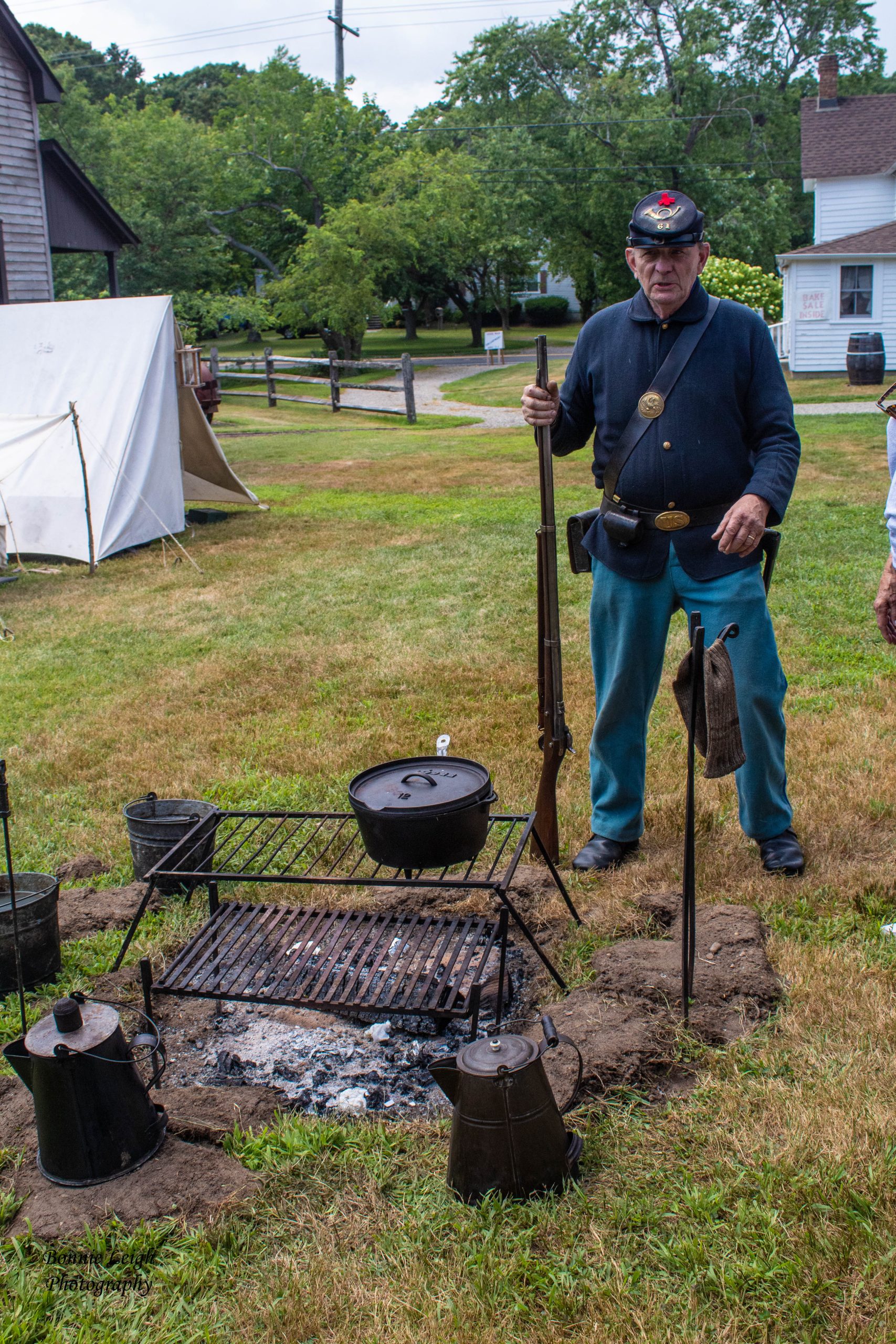 Photo of a man standing in front of a camp fire with pots and coffee pots around it and a tent in the background