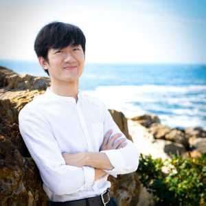 man in a white shirt standing against a rock on the beach with arms crossed