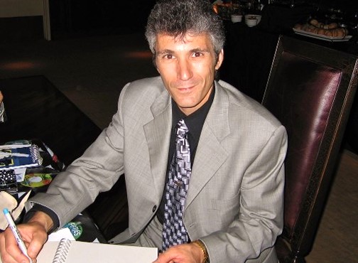 Man in a gray suit jacket and black shirt with grey tie sitting down writing