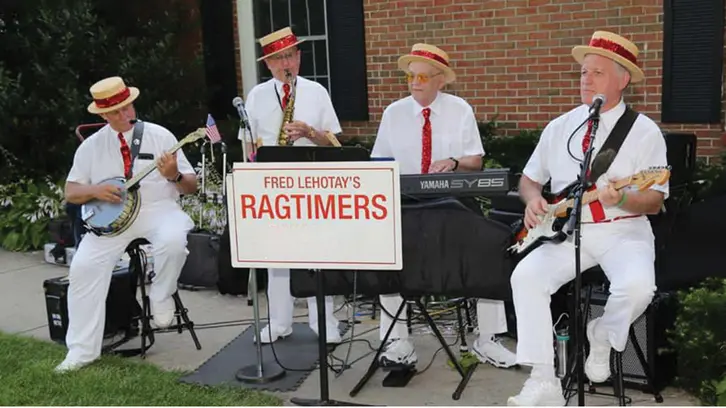 Four band members wearing all white with red ties and straw hats sitting outside on chairs playing music