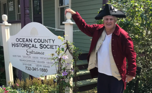 Man in a red coat white shirt and black hat leaning against a sign reading Ocean County Historical Society 